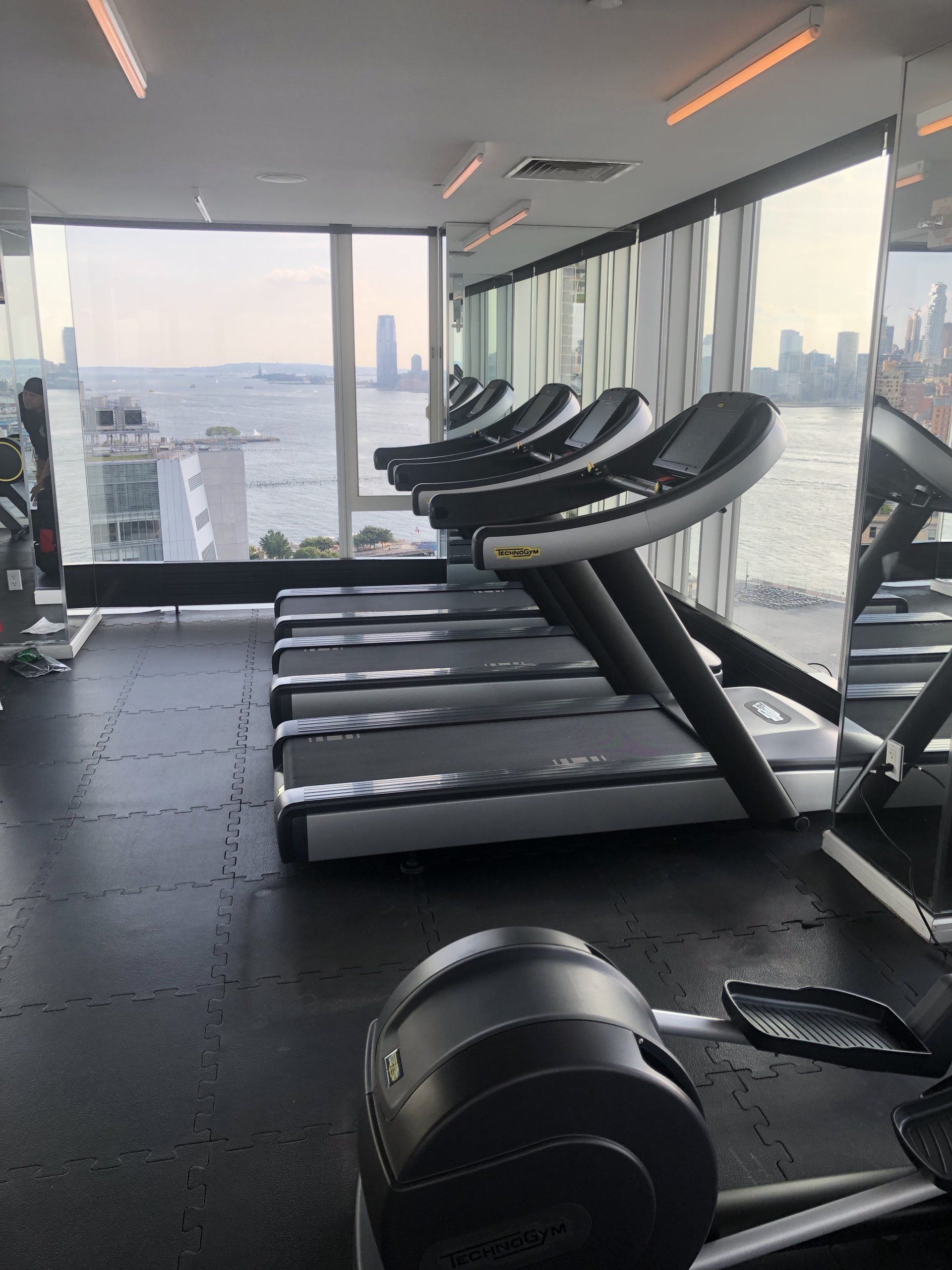 Row of treadmills in a high-rise gym with large windows overlooking city skyline.