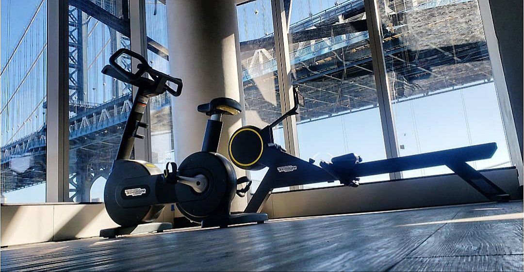 Stationary exercise bike and rowing machine positioned by large windows in a city gym.