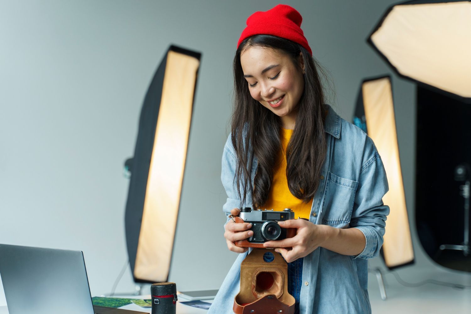 Woman with red beanie and denim shirt, holding a camera in a studio.
