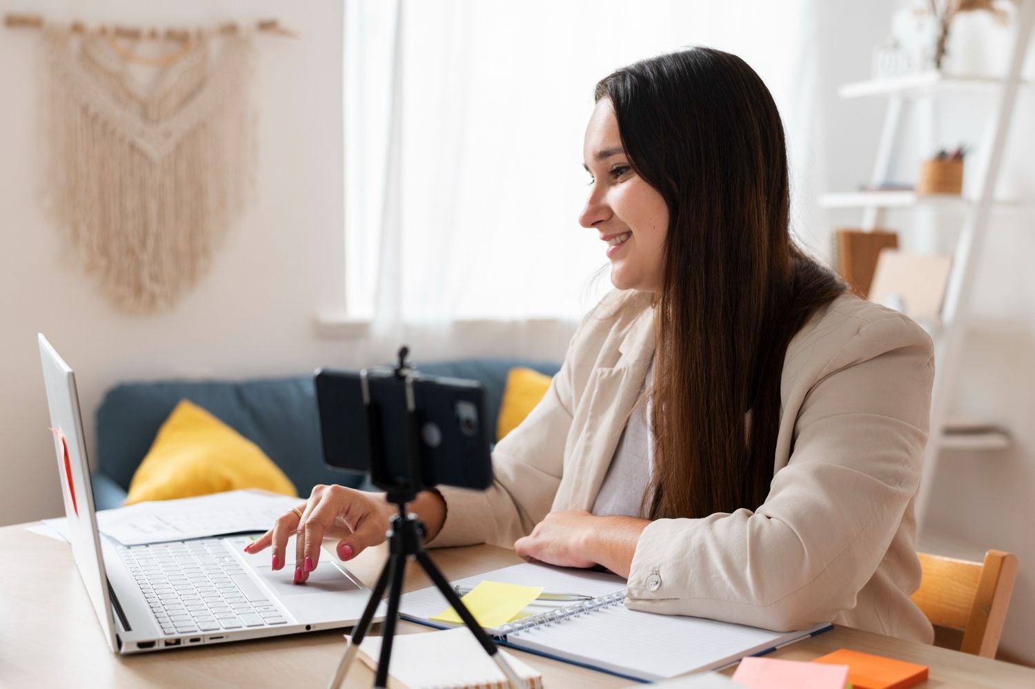Woman in a blazer works on a laptop, smiling. A phone on a tripod records, with a notebook and sticky notes visible.