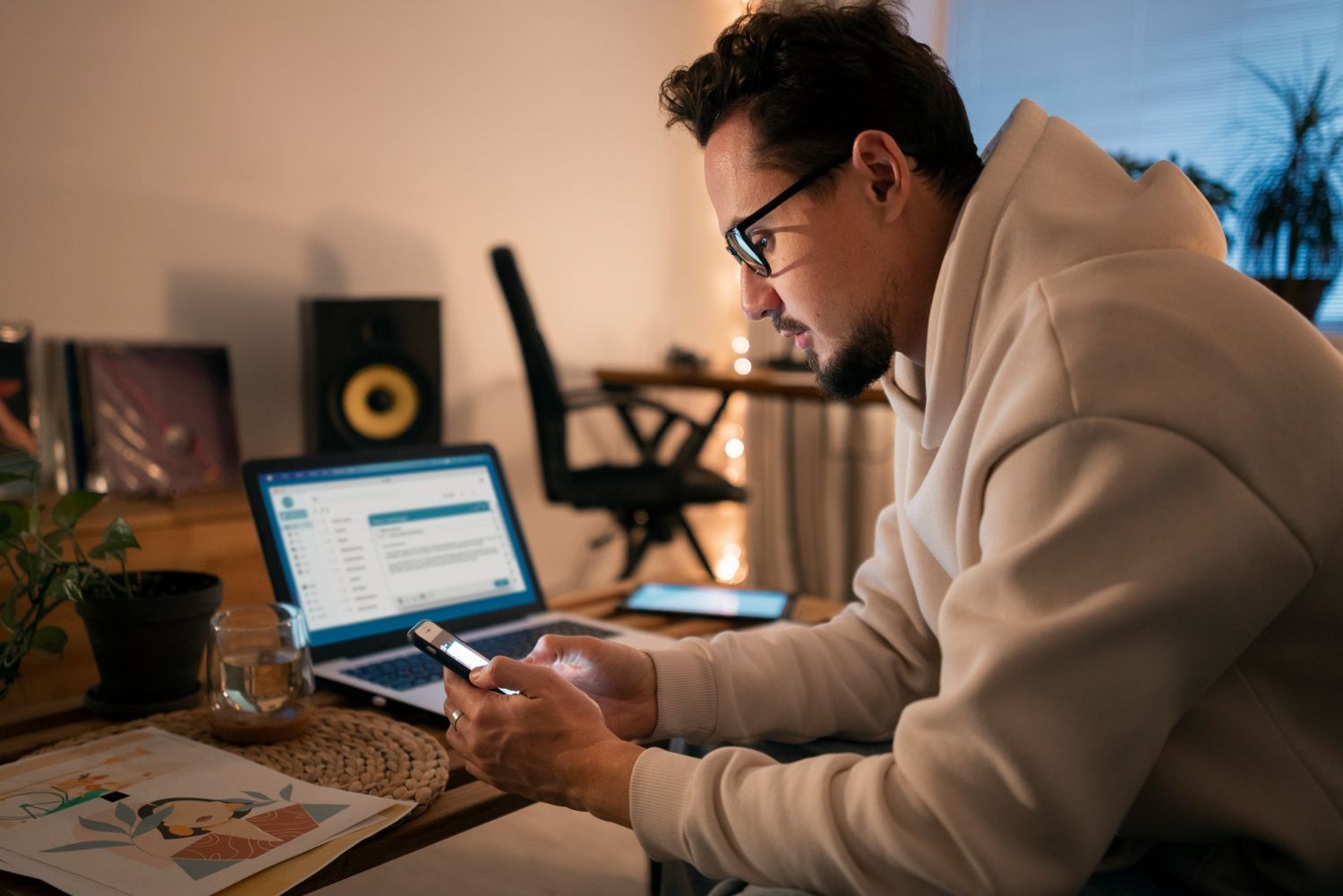 Man using laptop and phone in a dimly lit room, with books and speakers visible.