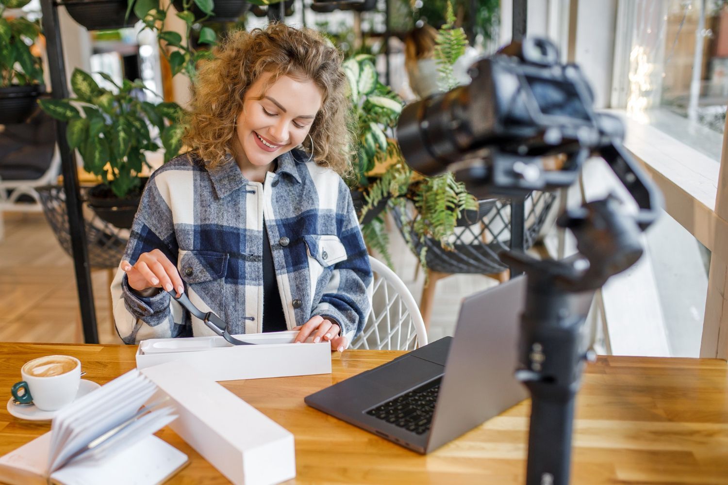 Woman filming unboxing video in a cafe, smiling at camera, laptop, camera, and food on the table.