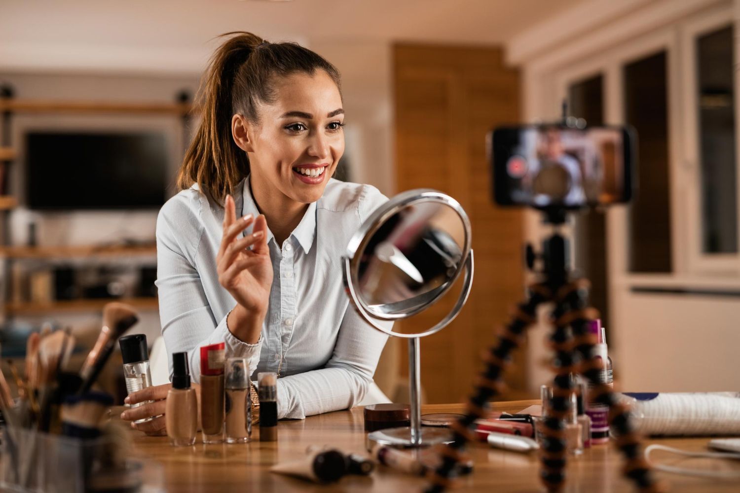 Woman filming makeup tutorial, smiling, with phone on tripod, mirror, and cosmetics.