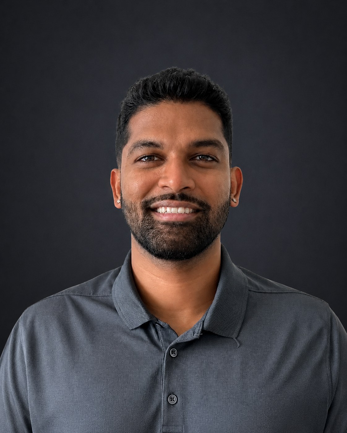 Man with dark hair and beard smiling, wearing a gray polo shirt against a white background.