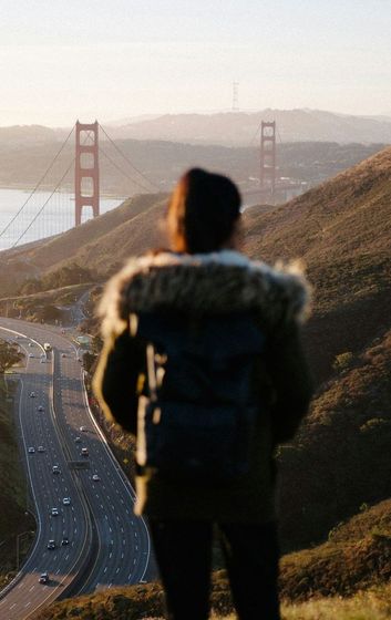 A woman is standing on top of a hill looking at the golden gate bridge.