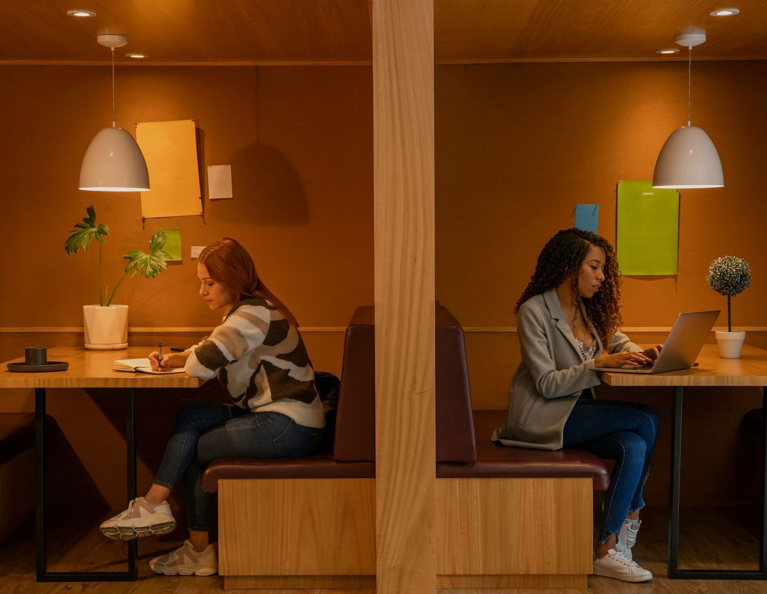 Two women are sitting at tables in a booth using laptops.