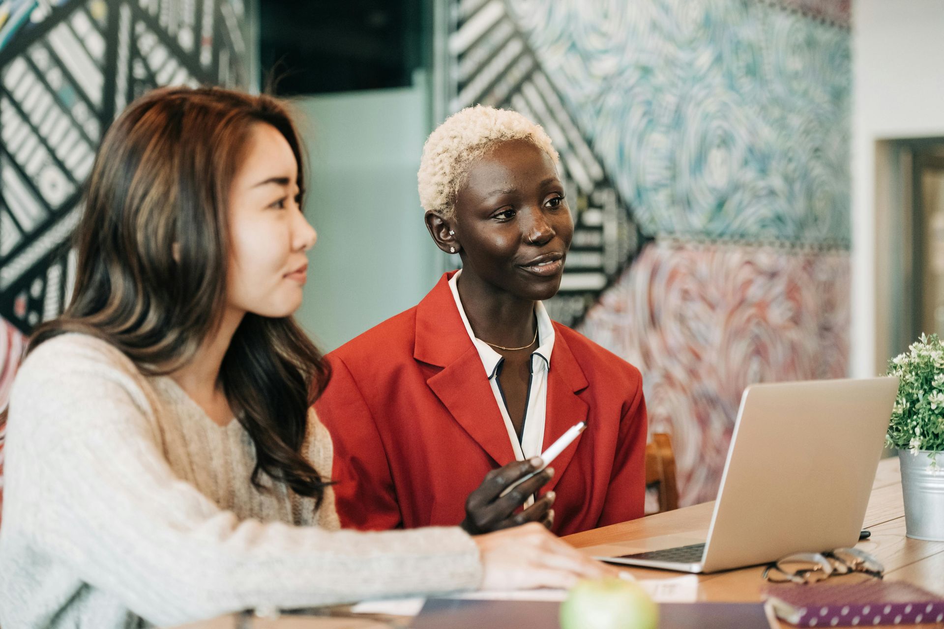 Two women are sitting at a table looking at a laptop computer.