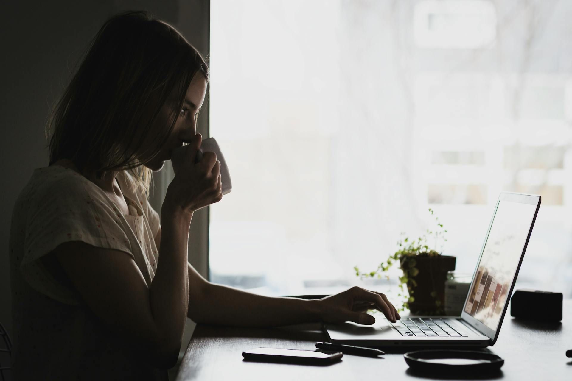 Woman sipping from a mug, working on a laptop near a window. Low light.