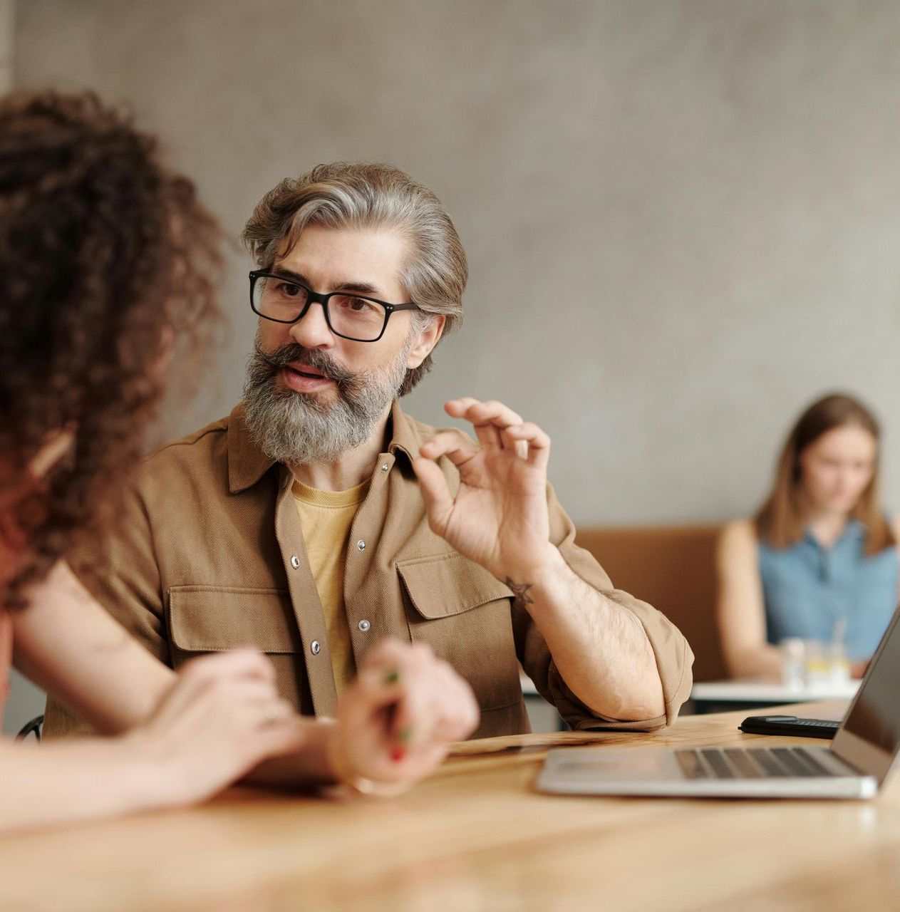 A man with a beard and glasses is sitting at a table talking to a woman.