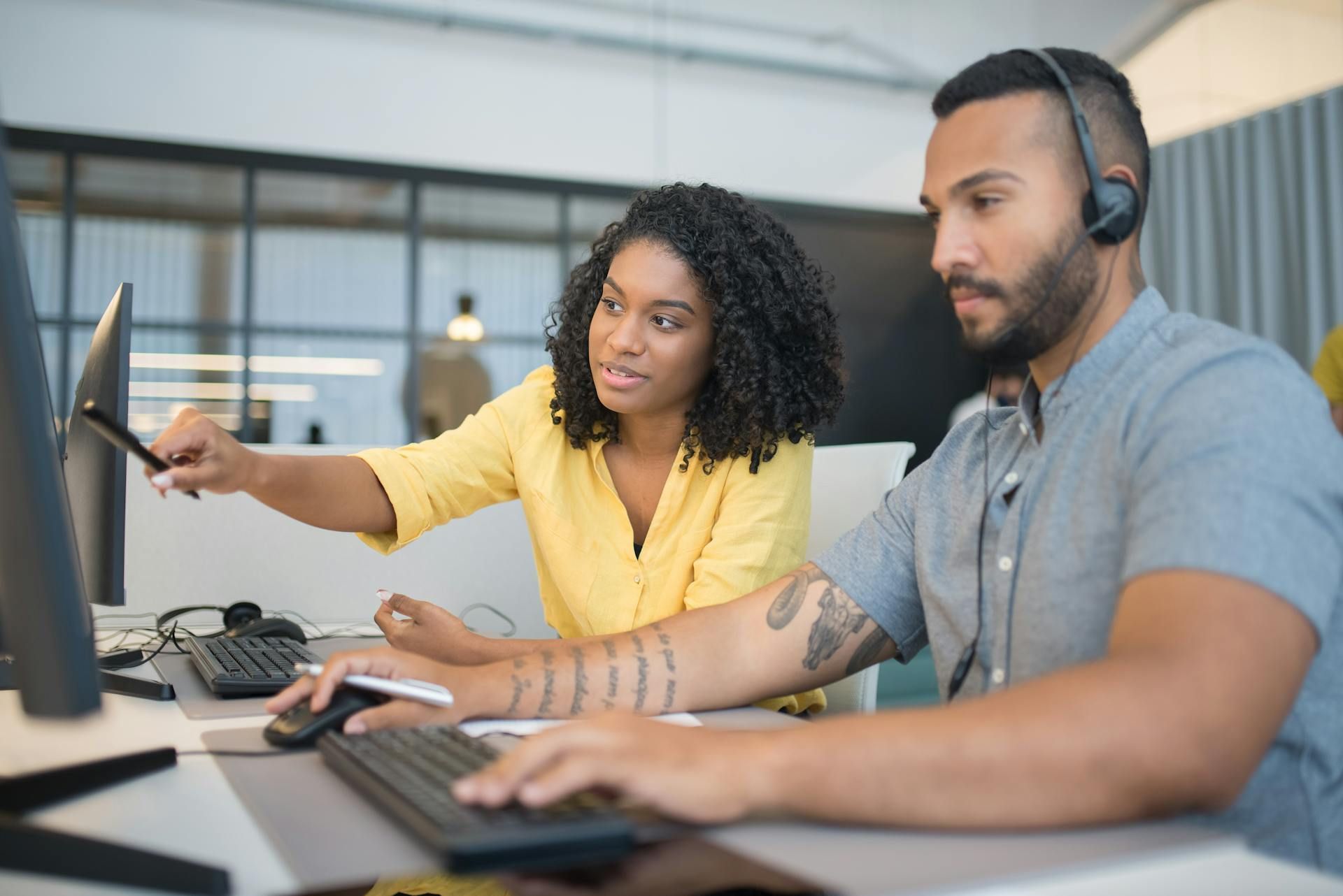 Woman pointing at a computer screen, assisting a man who wears a headset and is typing in an office.