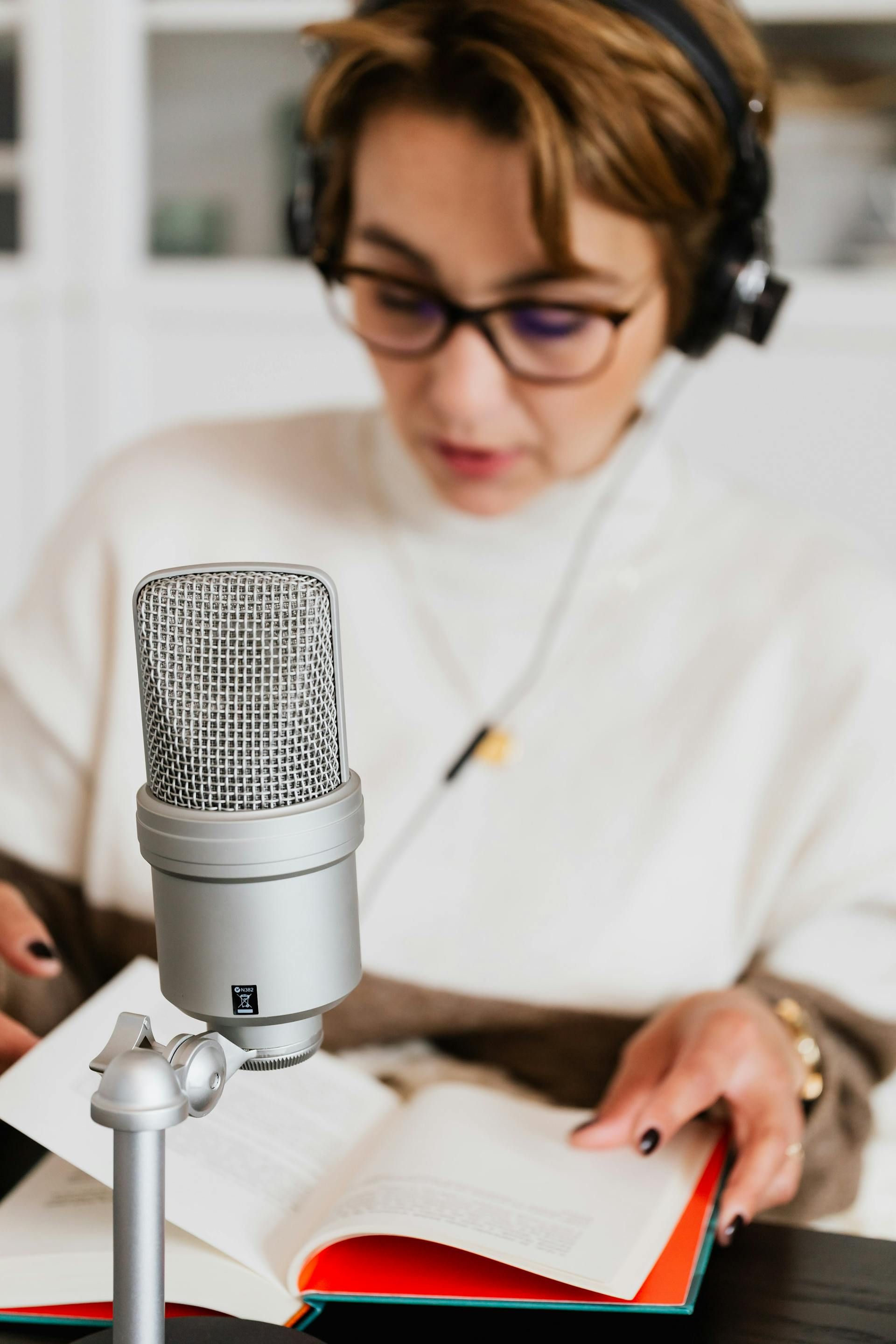 Woman with glasses and headphones reads into a microphone, sitting in front of a book, in a well-lit room.