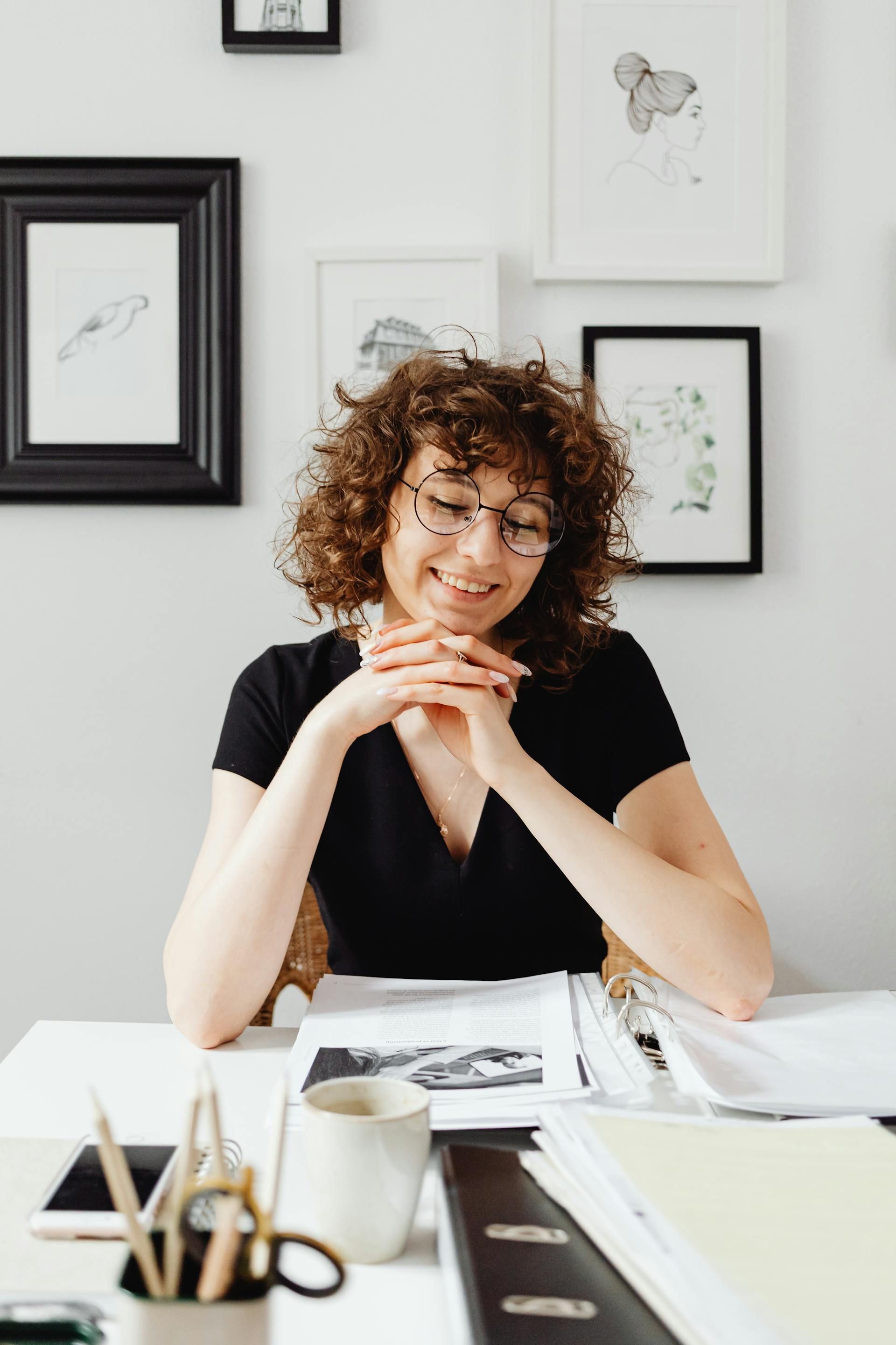 Woman with curly hair smiles, hands clasped, sits at a desk with papers, coffee, and pencils, framed art on the wall.