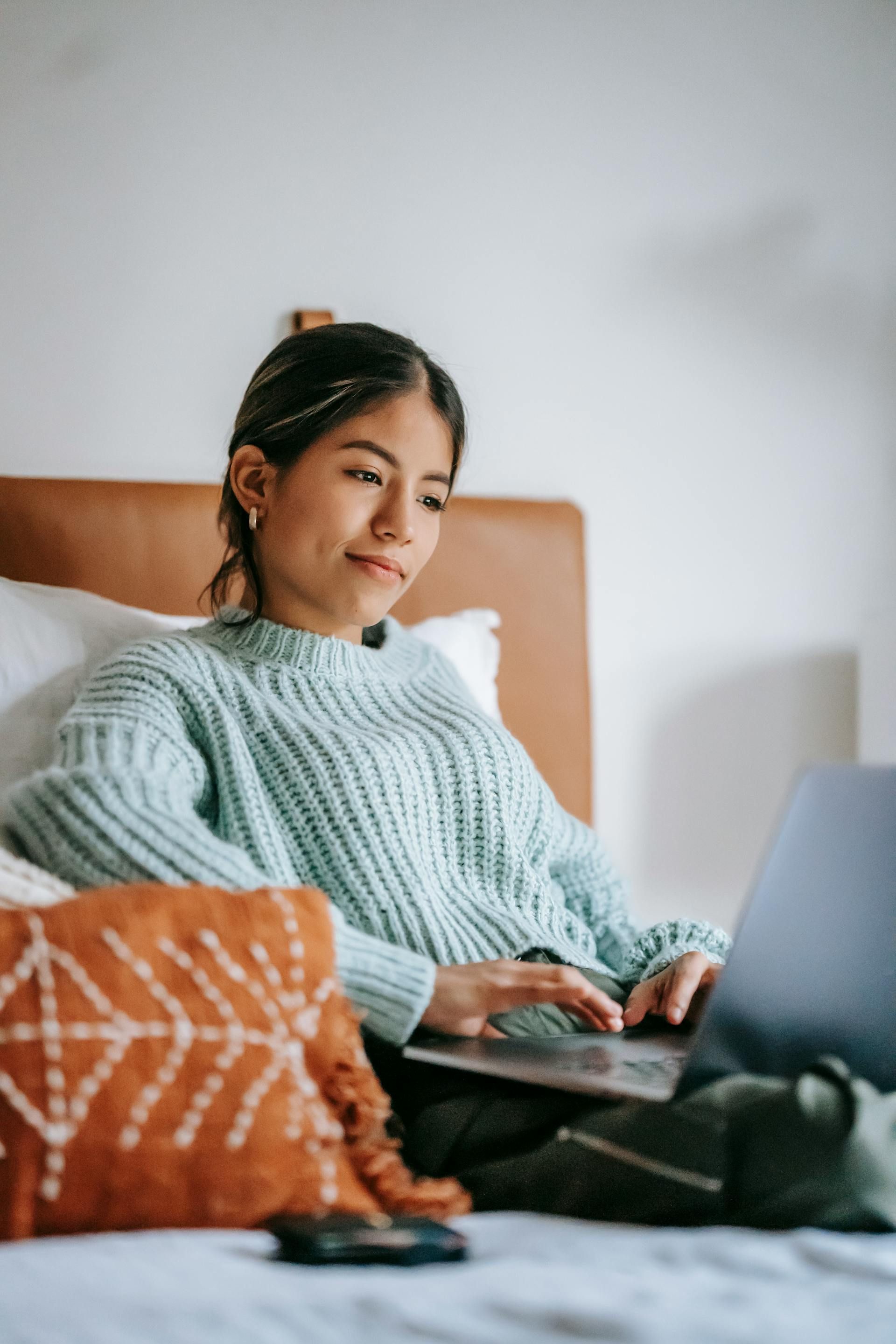 Woman sitting on a bed with a laptop, wearing a blue sweater, smiling.