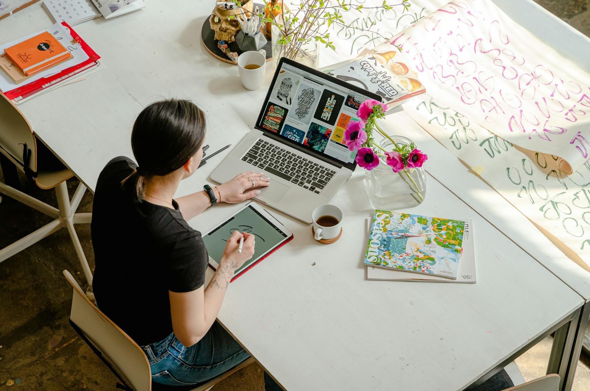 Person using a laptop and a tablet at a white table, flowers and coffee nearby.