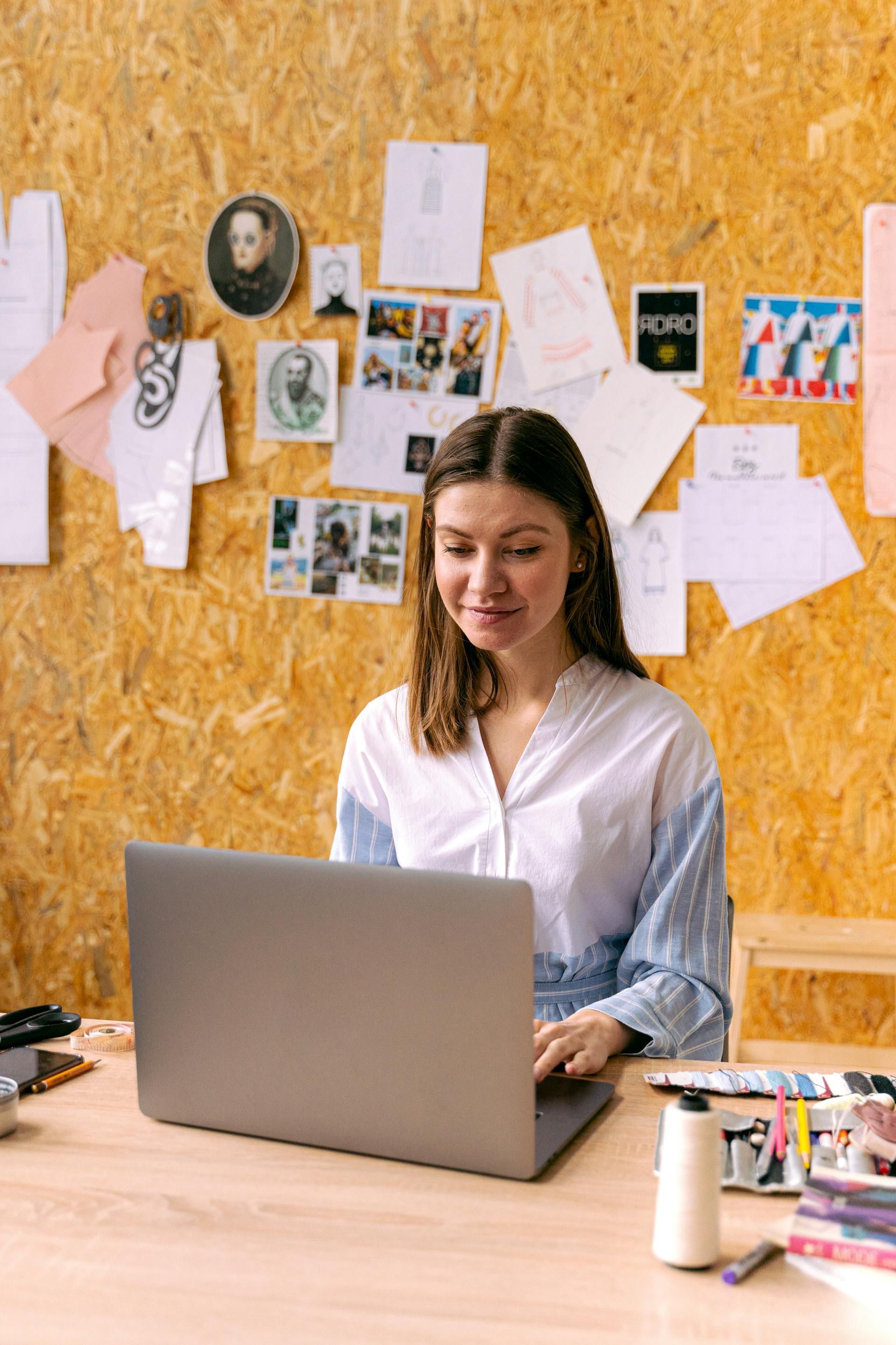 Woman working on laptop at a desk, smiling. Bulletin board with papers, photos, and sketches in the background.