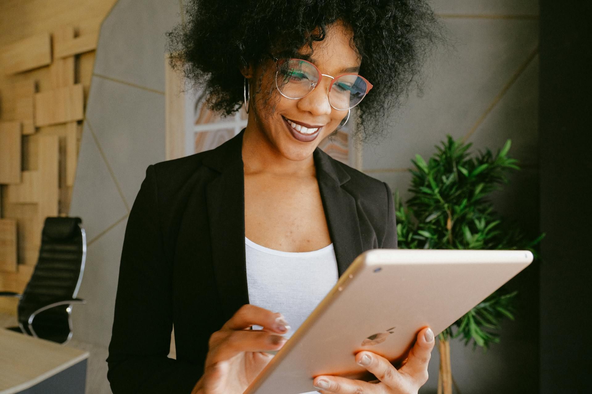Woman in blazer smiles, using a tablet indoors near a plant.