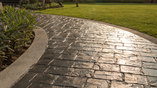 Curved brick-patterned walkway with a concrete border, leading through a grassy yard with plants.