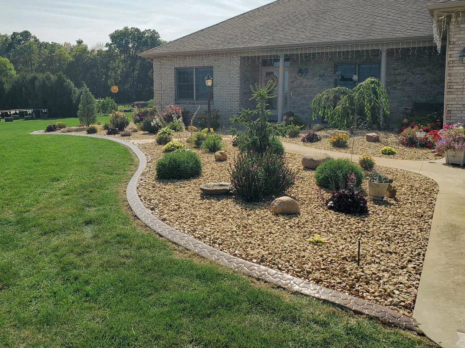 House with landscaping; tan gravel bed, green lawn, various plants, and a concrete border.