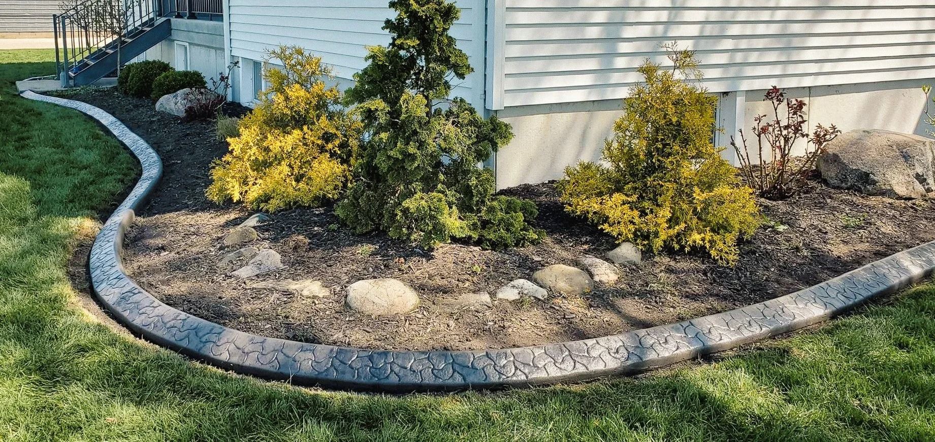 A landscaped flower bed with dark gray concrete edging, shrubs, and mulch beside a light-colored house.
