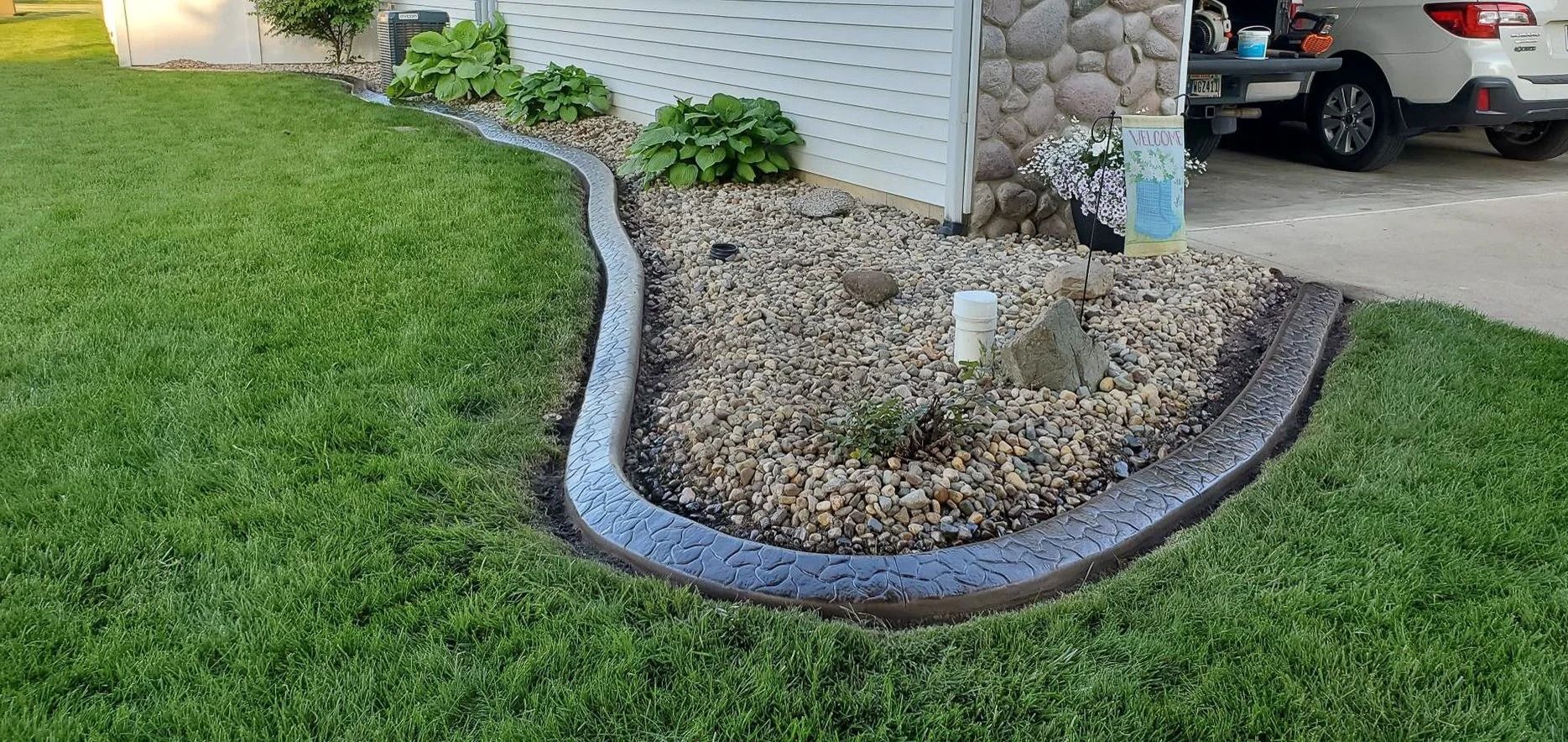 Lawn with concrete border edging a rock garden next to a house with a car parked nearby.