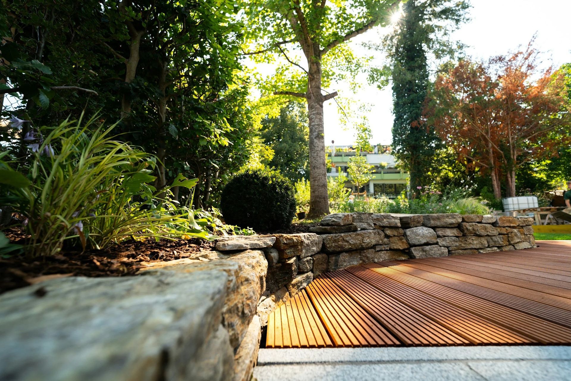 Stone wall and wooden deck in a garden with lush greenery and trees. Sunlight streams through.
