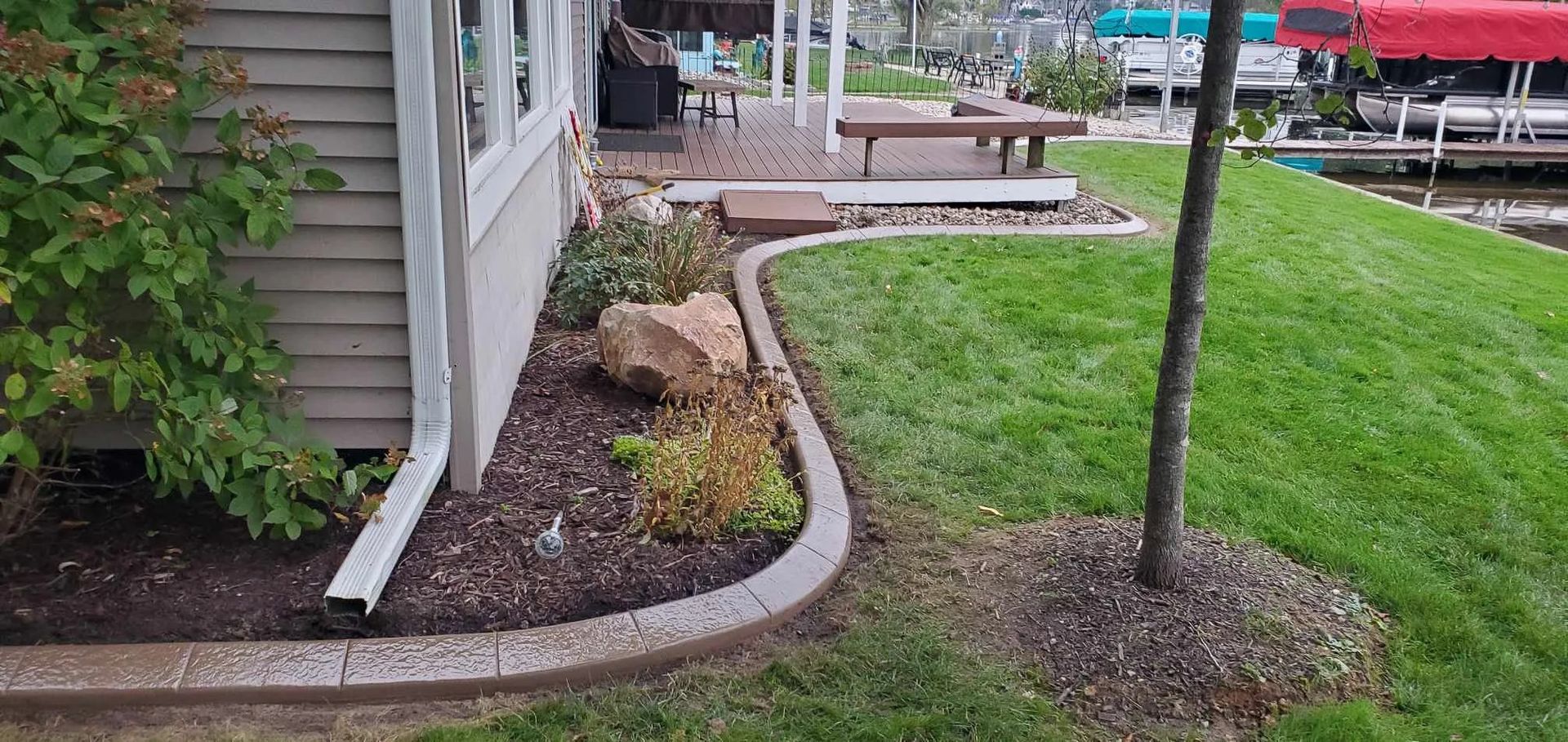 Lawn next to house with concrete edging. Mulch, plants, and a tree are present. Lake in the background.