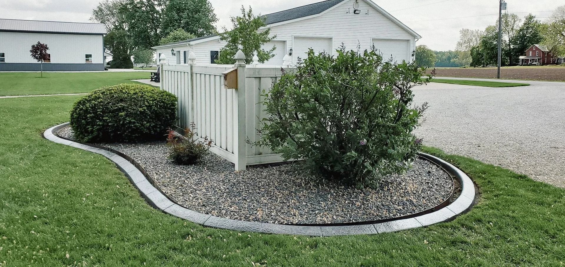 A landscaped garden bed with a white fence, surrounded by green grass and a gravel driveway.