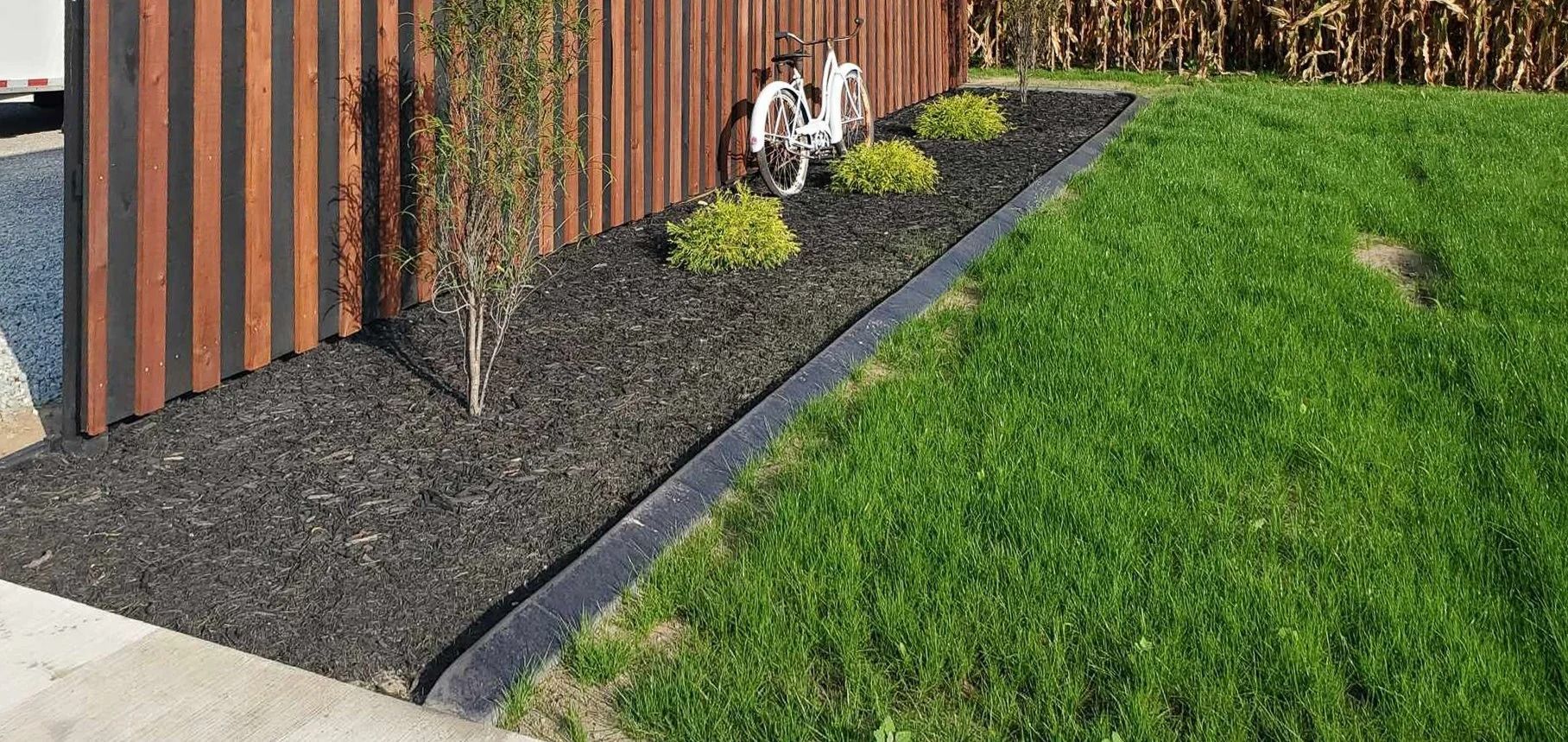 Landscape bed with dark mulch, edged by black border, next to green lawn and brown fence.