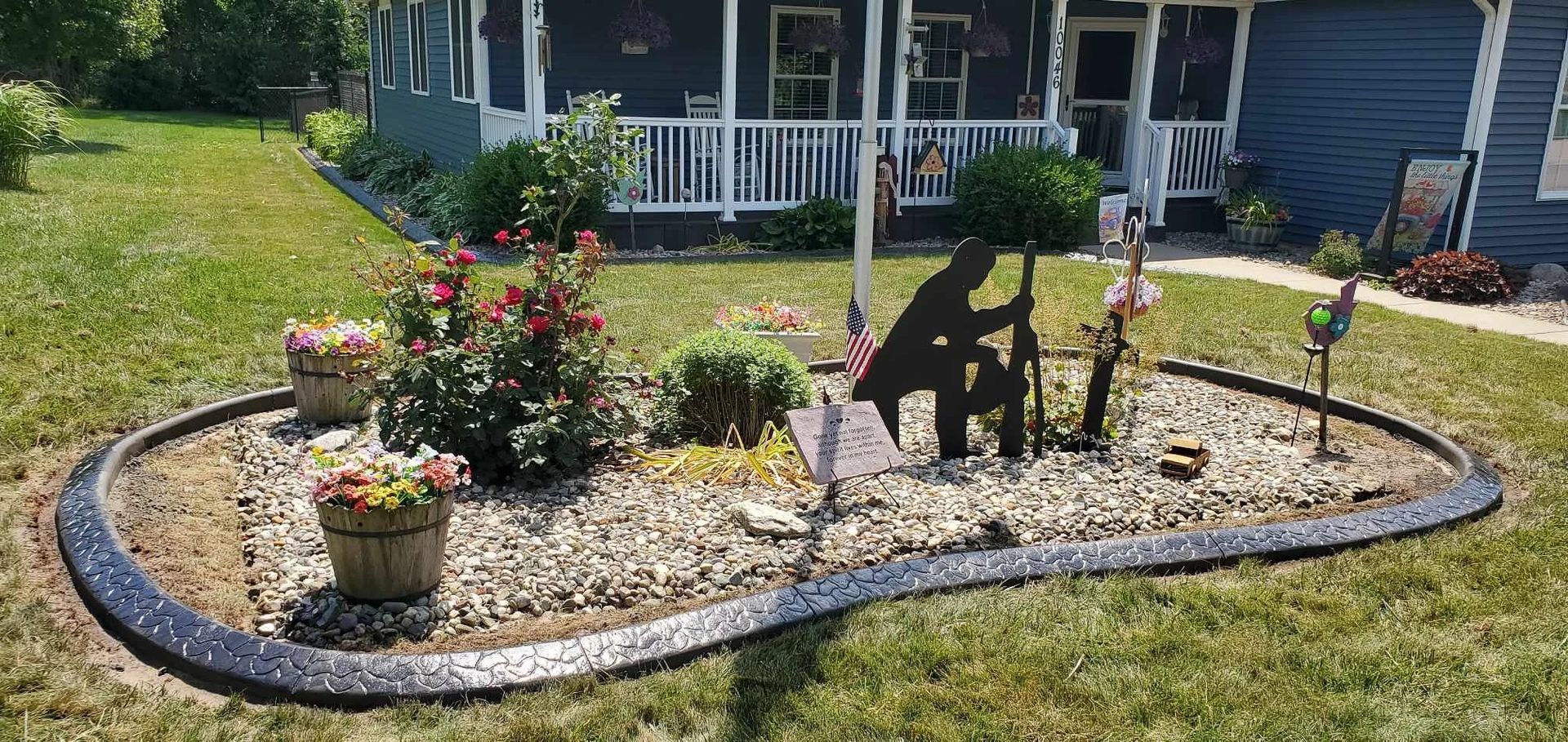 A front yard flower bed with a memorial statue and potted flowers, bordered by concrete.