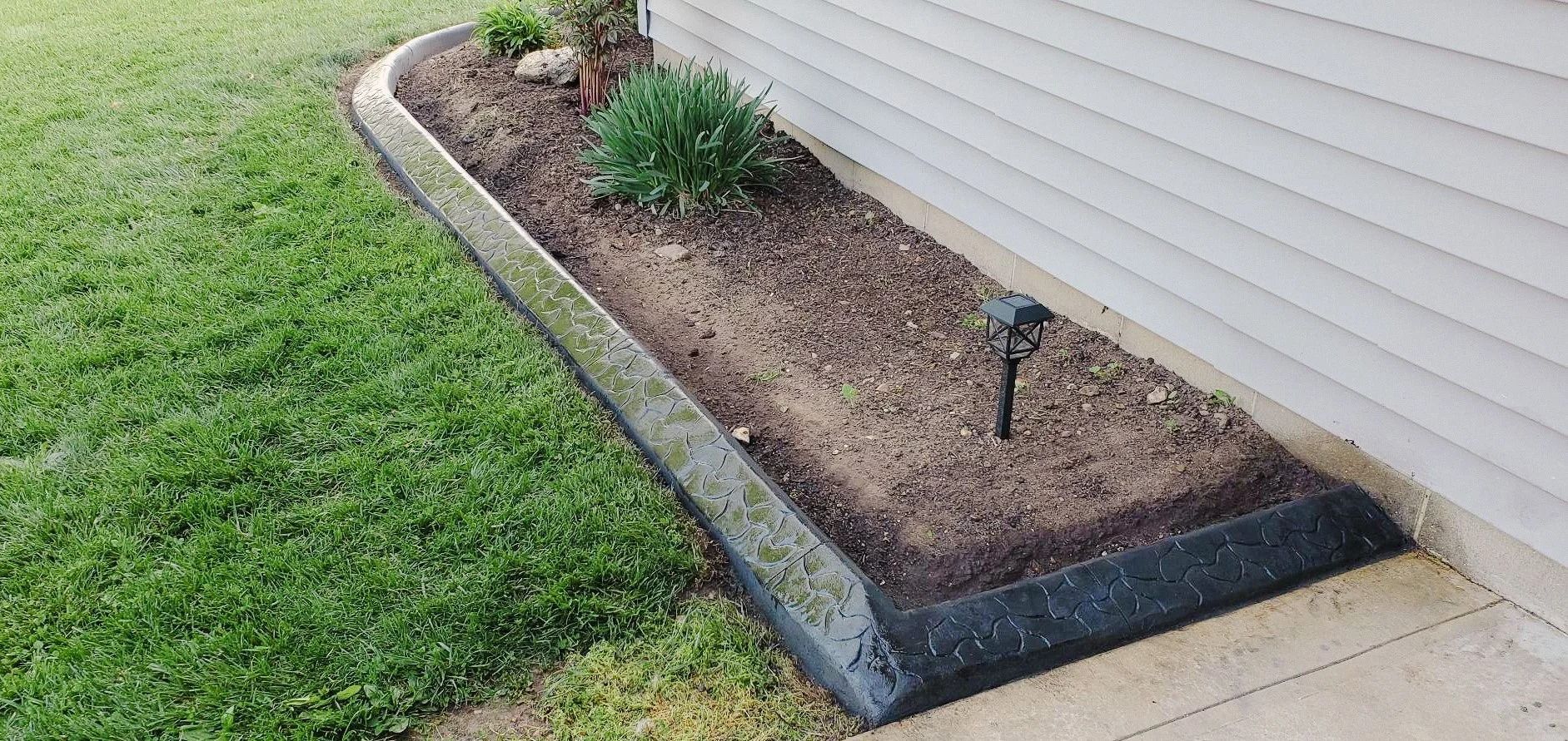 A raised garden bed with mulch and plants next to a house and grass.