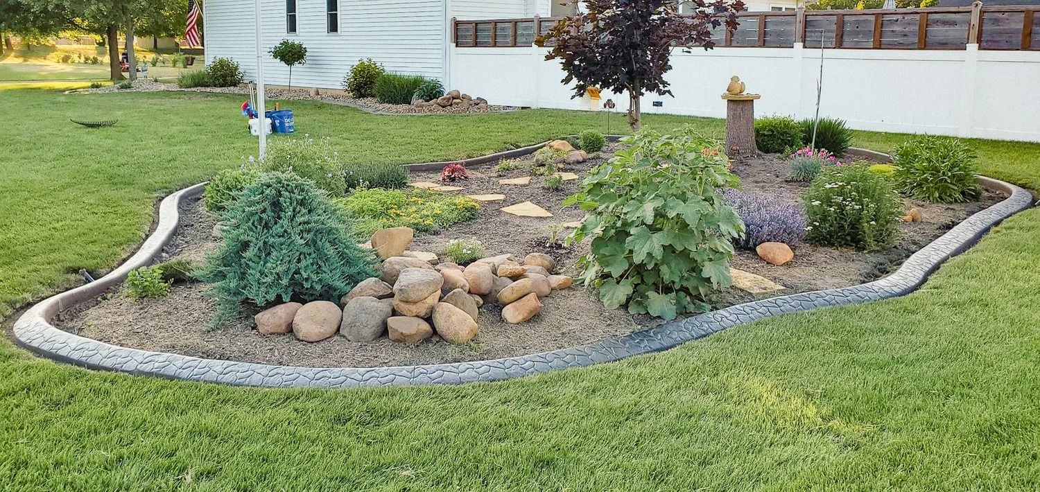 Garden bed with gray stone edging, various plants, and rocks on a green lawn.