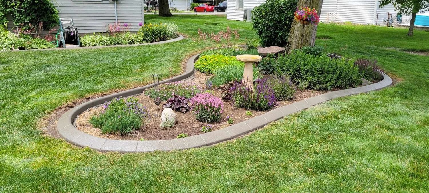A garden bed with a concrete border surrounded by grass, featuring flowers, a bird bath, and a small animal.