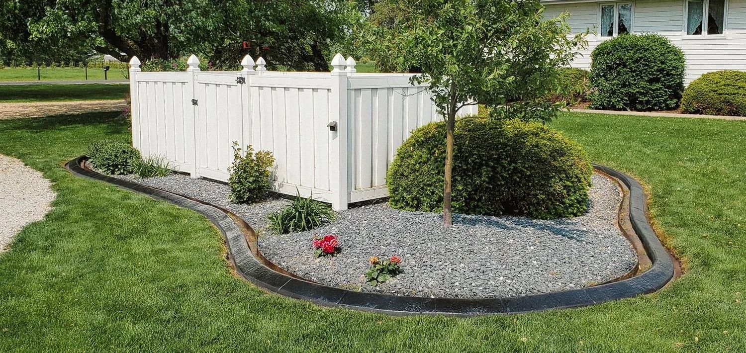 White picket fence surrounds a landscaped area with mulch, plants, and a small tree.