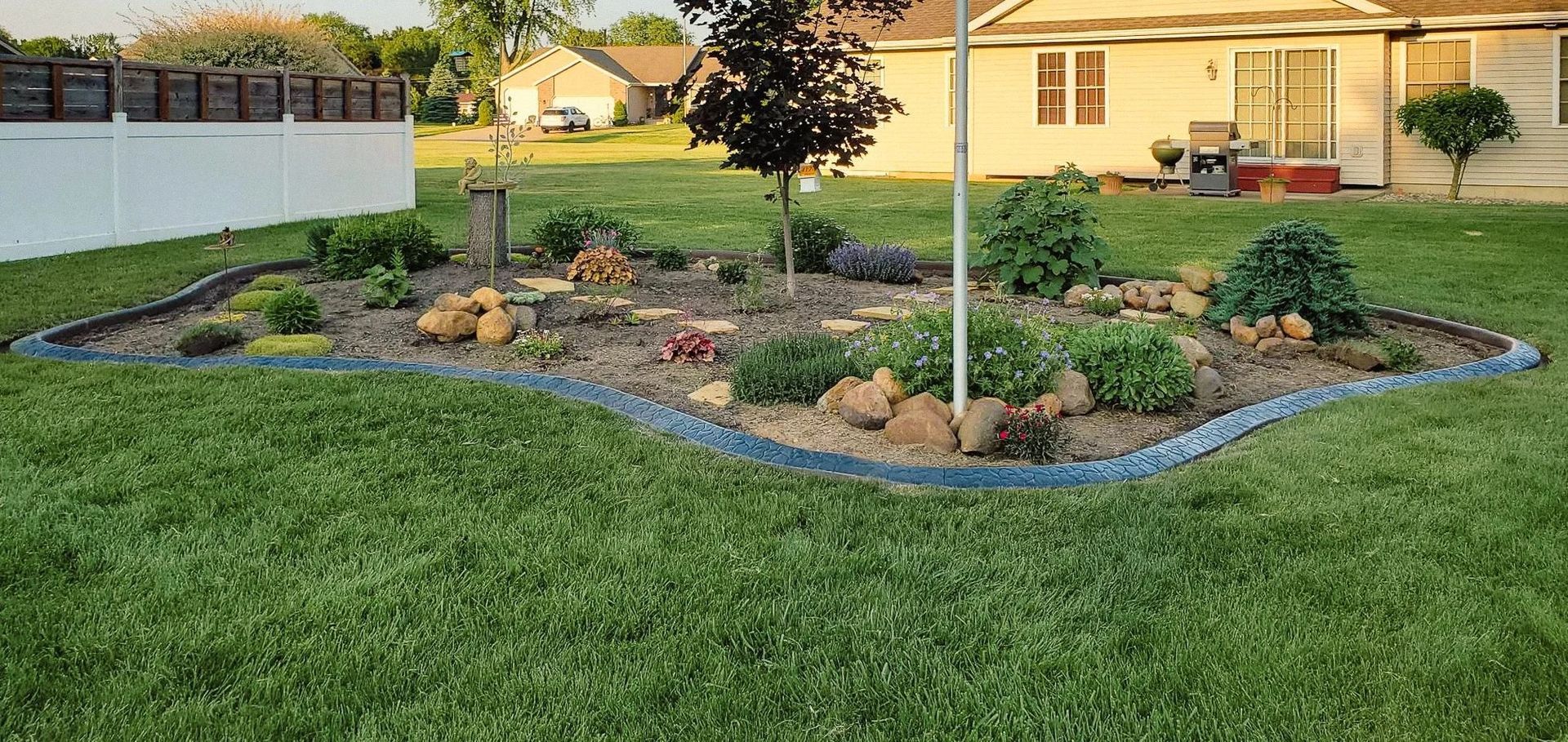 A backyard garden with dark edging, green grass, and a house in the background.