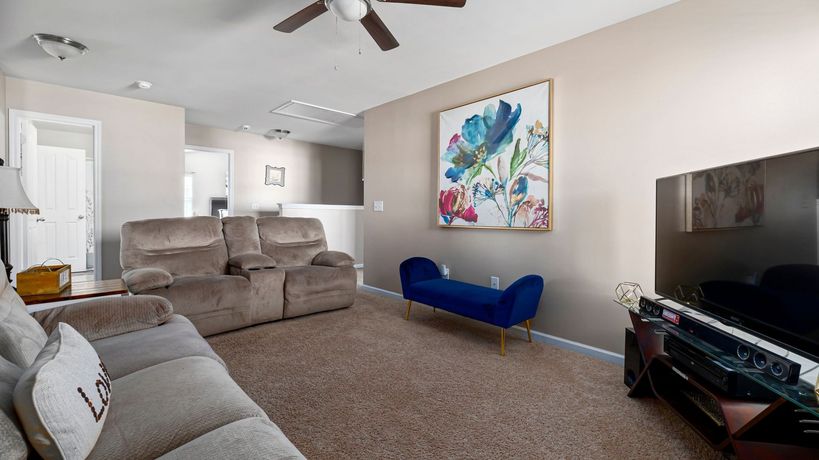 Living room with beige couch, TV, and floral art, blue bench, and brown ceiling fan. Living room with beige couch, TV, and floral art, blue bench, and brown ceiling fan.