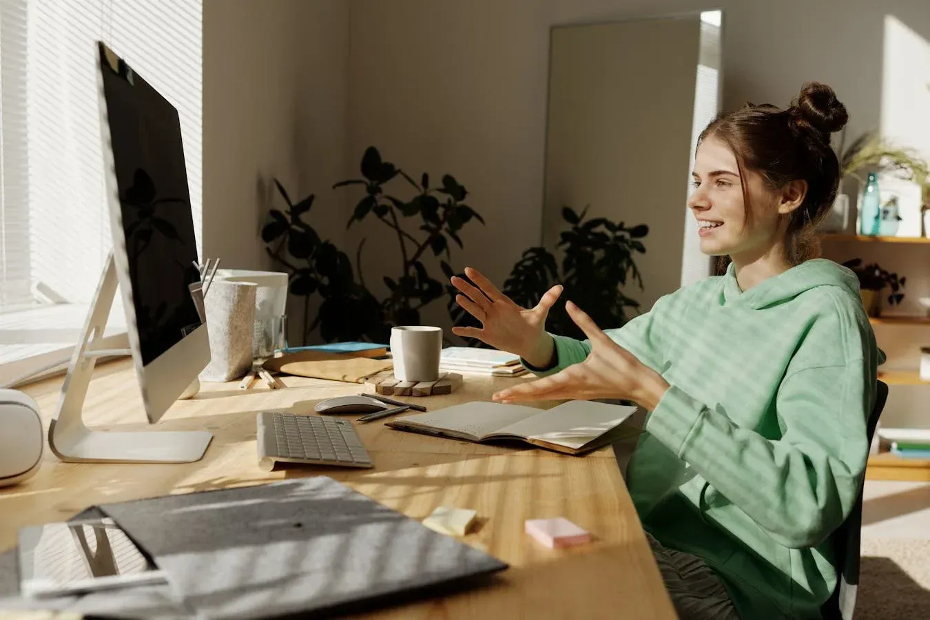 Woman gesturing while on a video call at a desk with a computer and notebook. Sunlight streams in.