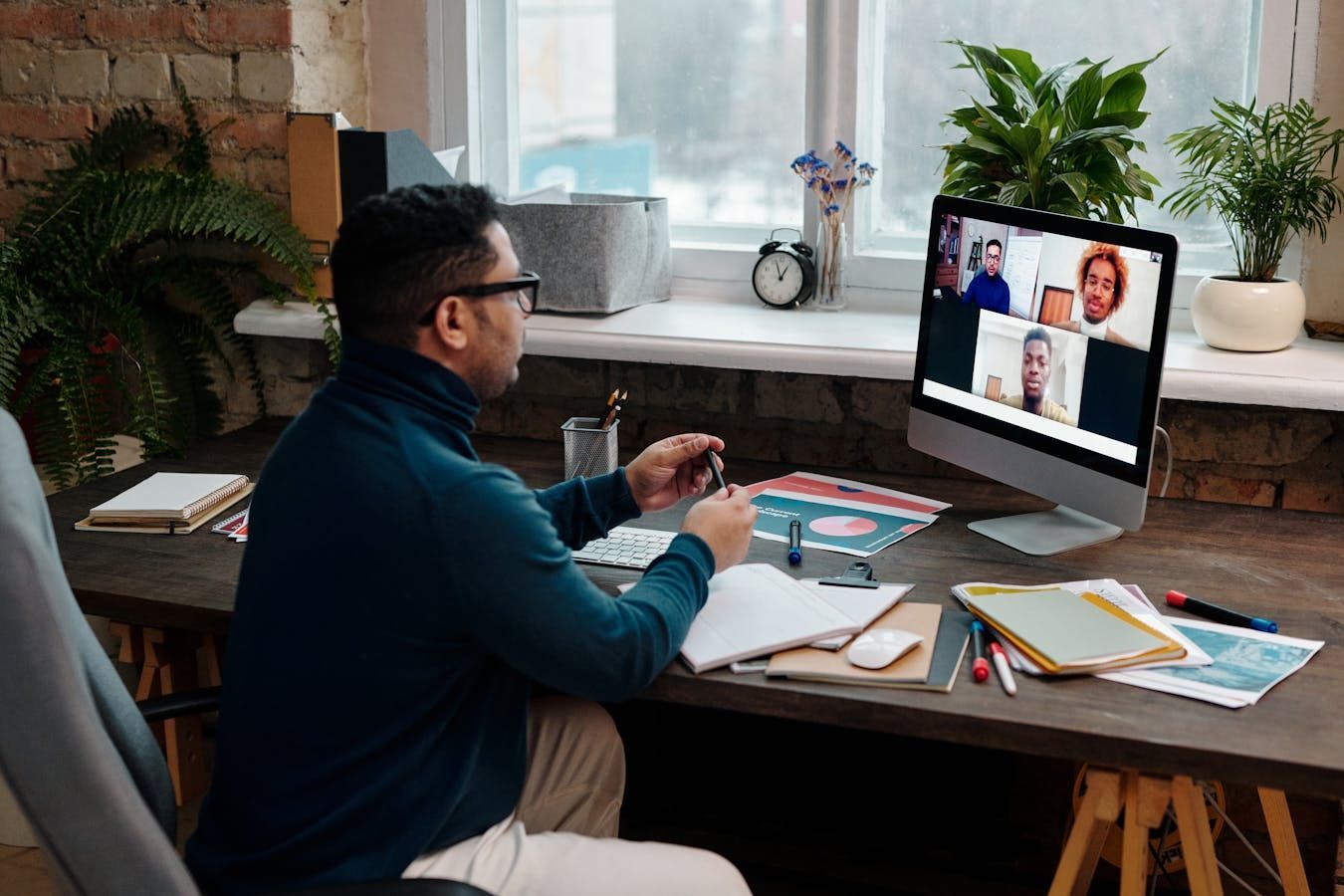 Man in a blue sweater at desk having video call with colleagues. Desk has notes, plants, and a computer.