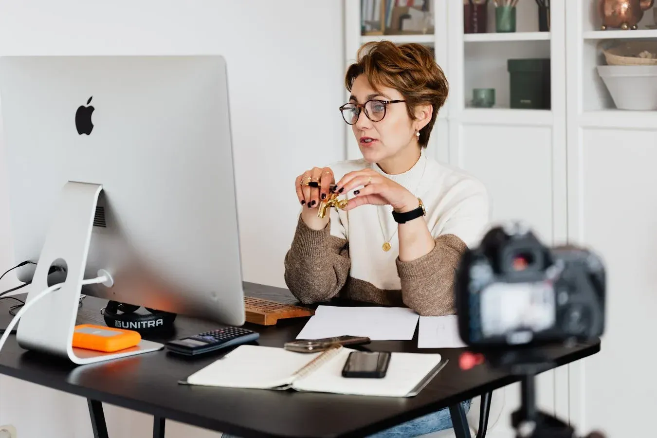 Woman with glasses at a desk, looking at a computer monitor and speaking; camera in foreground.