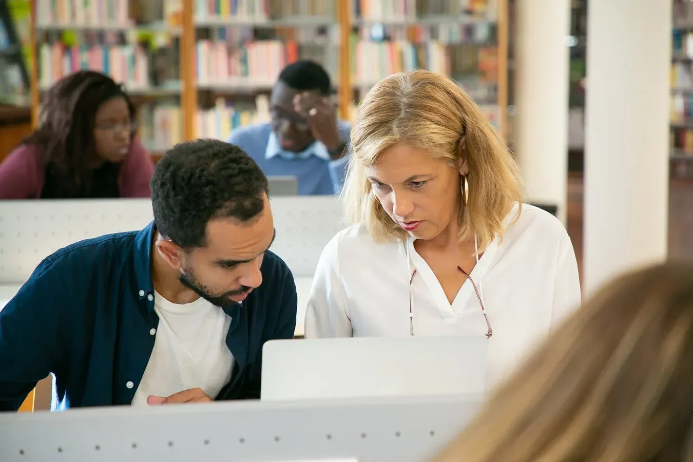 Two people looking at laptop in library, surrounded by others reading.