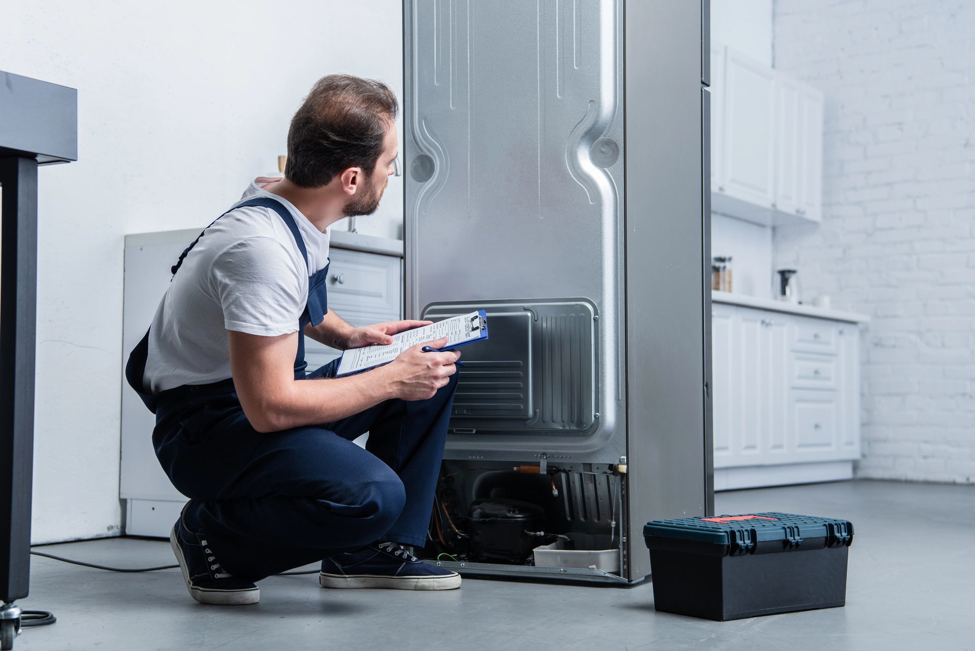 Un técnico se arrodilla, inspeccionando la parte trasera de un refrigerador en una cocina