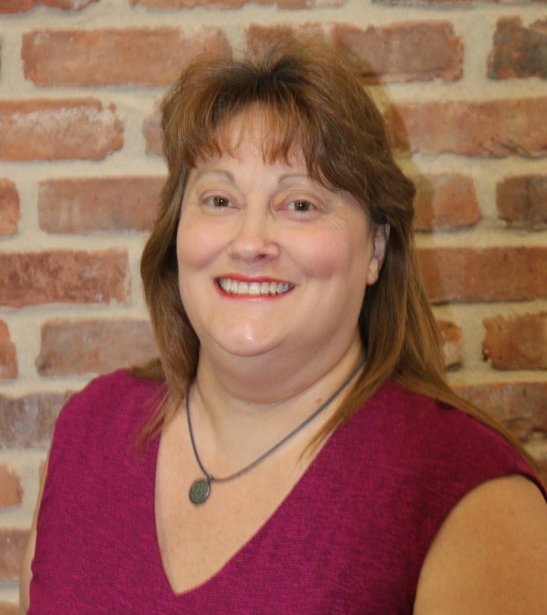 A woman in a purple shirt is smiling in front of a brick wall.