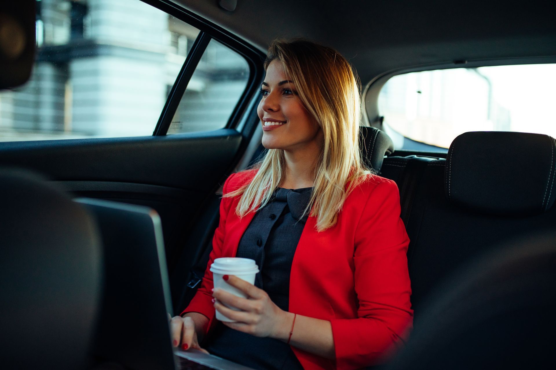 Woman in red blazer in car, smiling, holding coffee, with laptop open.