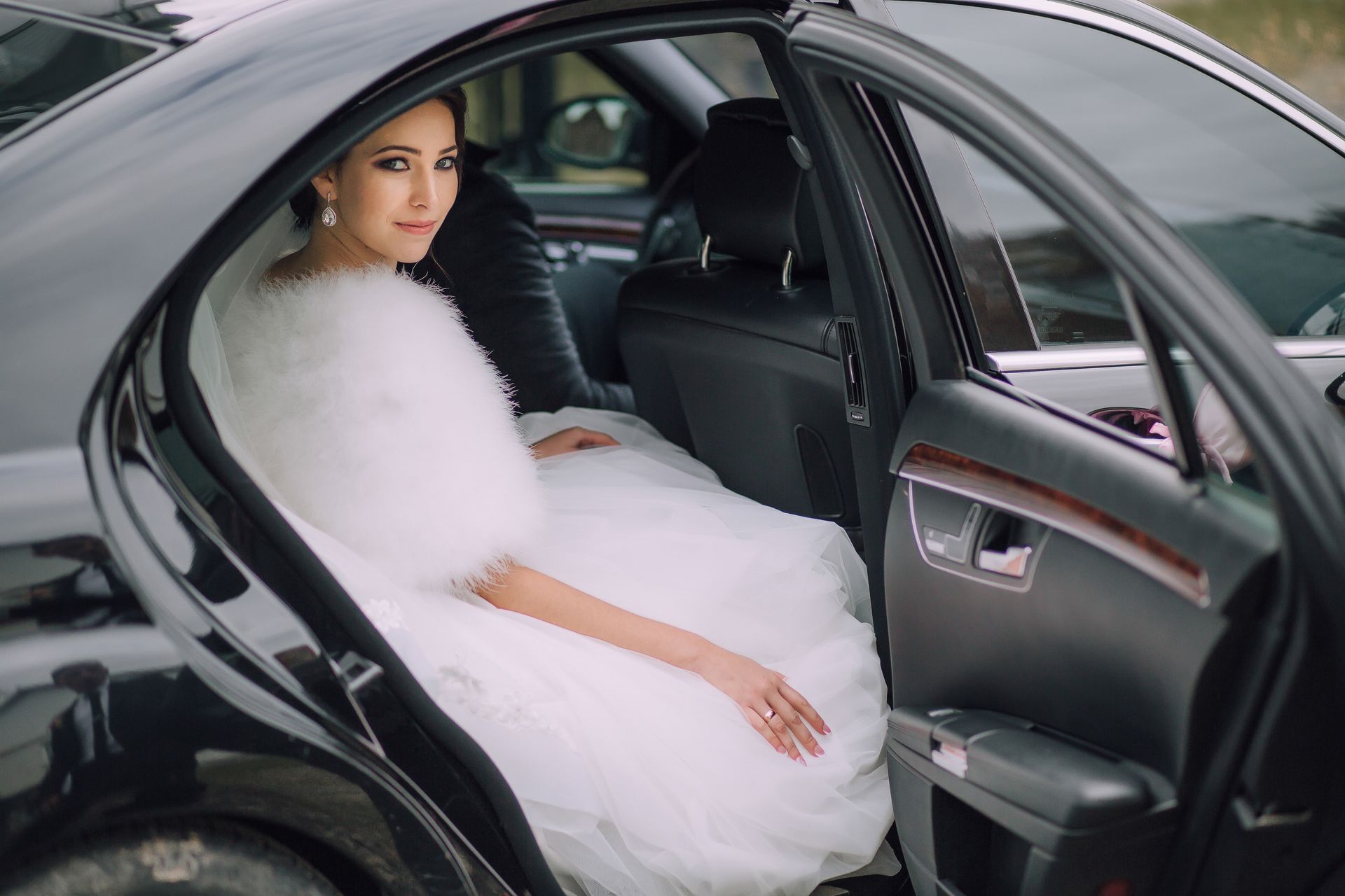 Bride in a white dress and fur shawl sits in a black car, smiling.