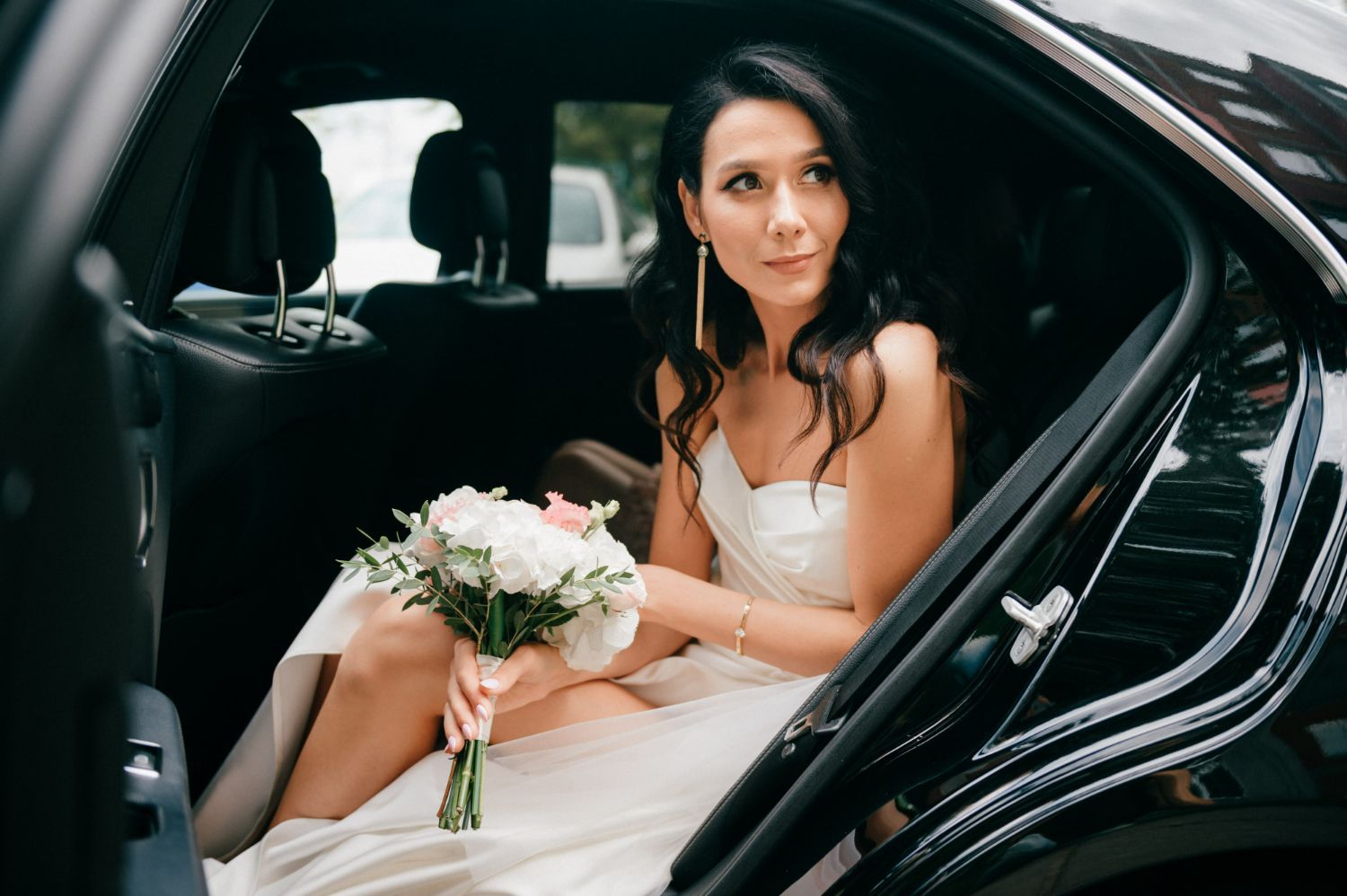 A bride is sitting in the back seat of a car holding a bouquet of flowers.