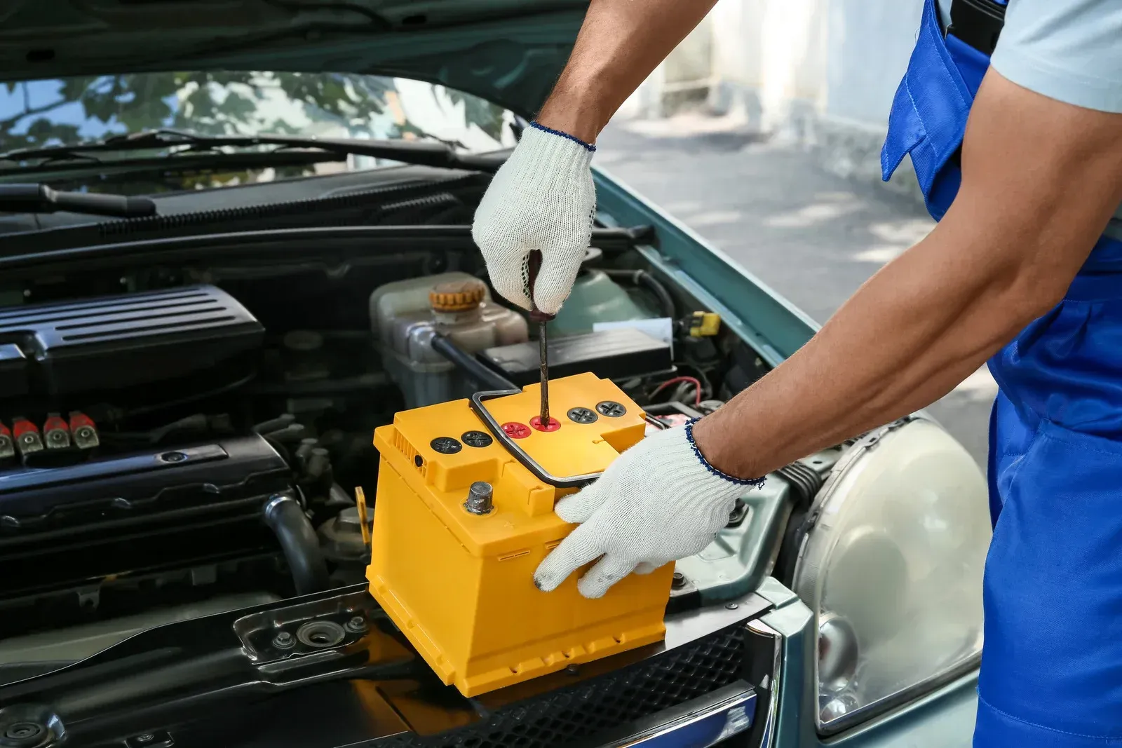 Mechanic in blue overalls and gloves working on a car battery in an engine bay.