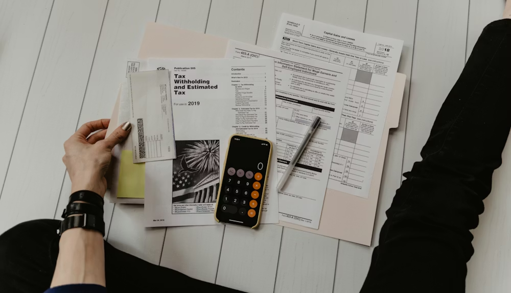 Hands holding papers beside a calculator and smartphone on a white desk with charts and notes
