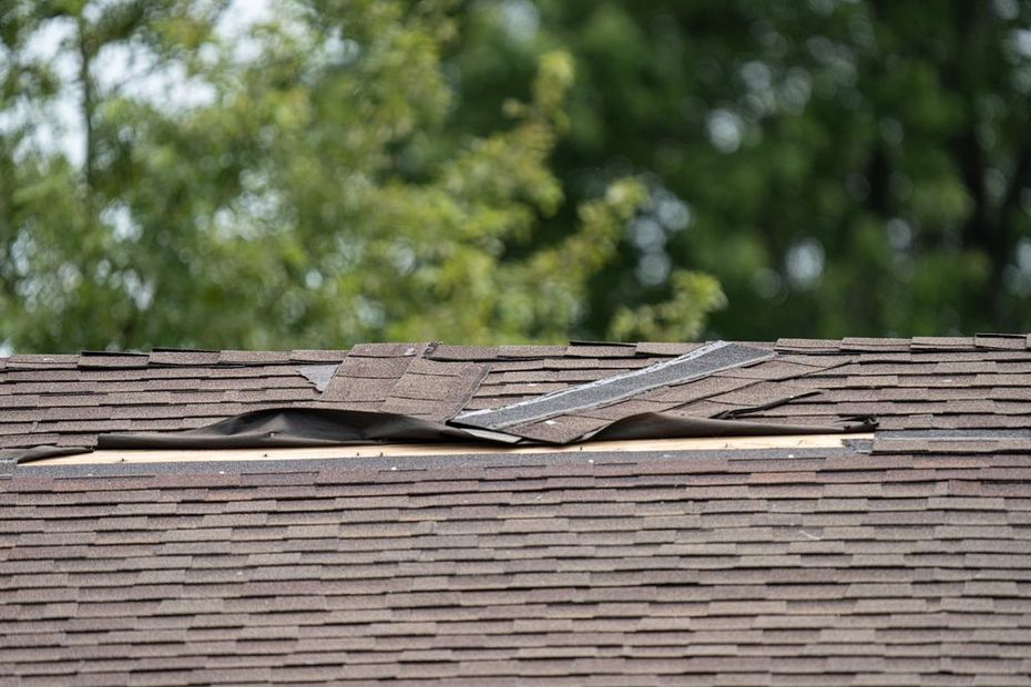 Damaged shingled roof with a large hole and torn sections against a blurred tree background