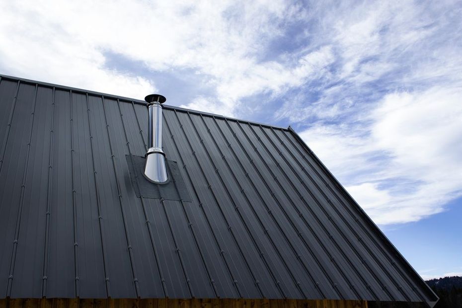 Dark metal roof with a silver vent pipe against a cloudy blue sky