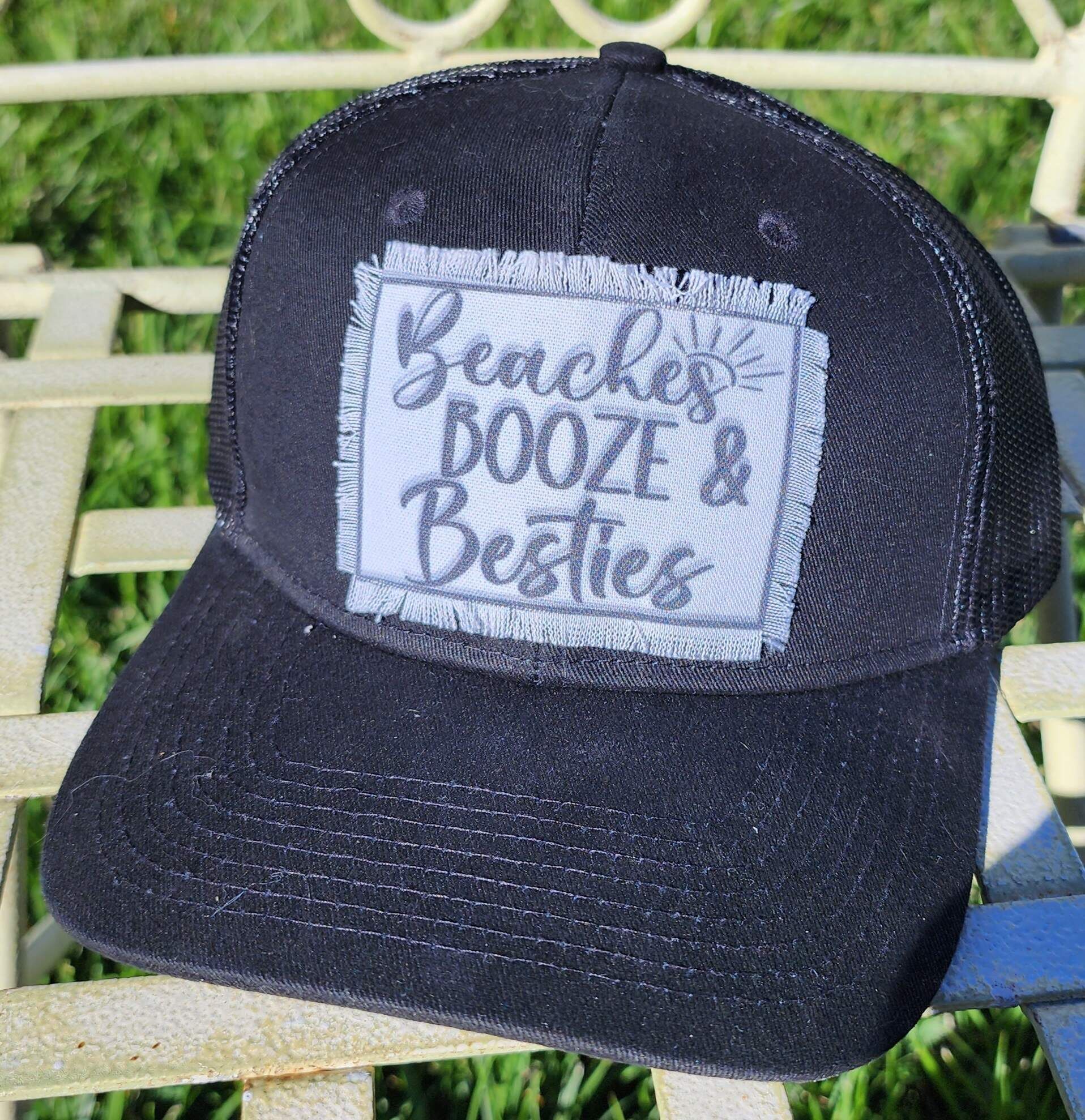 A black trucker hat with the words `` beaches dooze & besties '' embroidered on it is sitting on a chair.