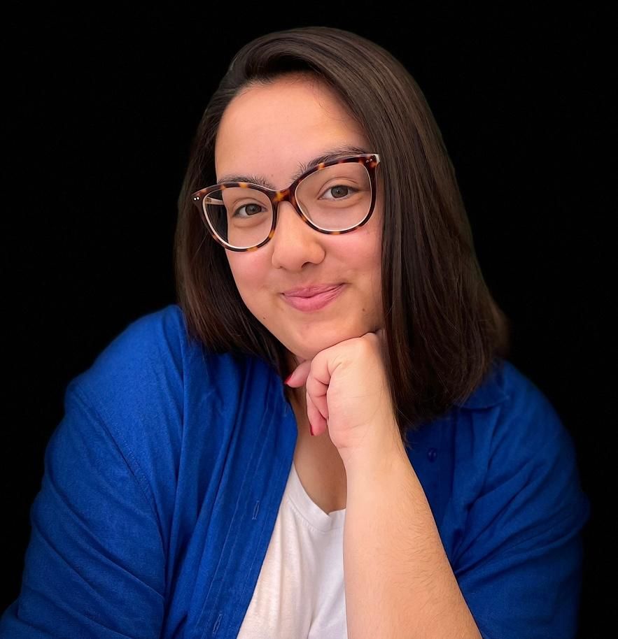 Woman with glasses, resting chin on hand, smiles, wearing blue sweater, white shirt, dark background.